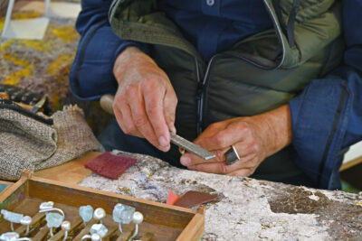 zwei Hände arbeiten mit einer Feile an einem Stück Metall, im Vordergrund ein Kasten mit Ringen